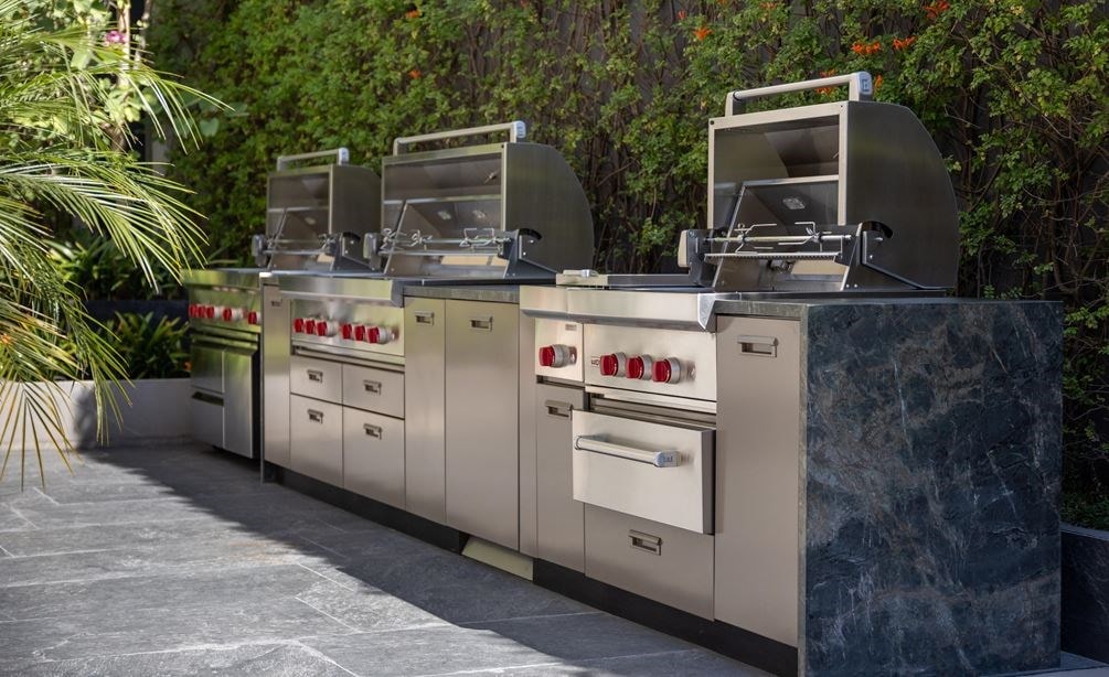 Three outdoor grills side-by-side a part of Wolf Appliances in front of a brush background.
