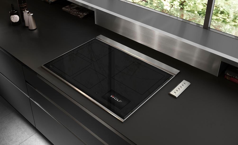 Placa de inducción transicional de 30" Wolf (CI30460T/S) exhibida en un elegante diseño de cocina ultramoderno con mostradores negros lisos, salpicadero de acero inoxidable y gabinetes sin manija 