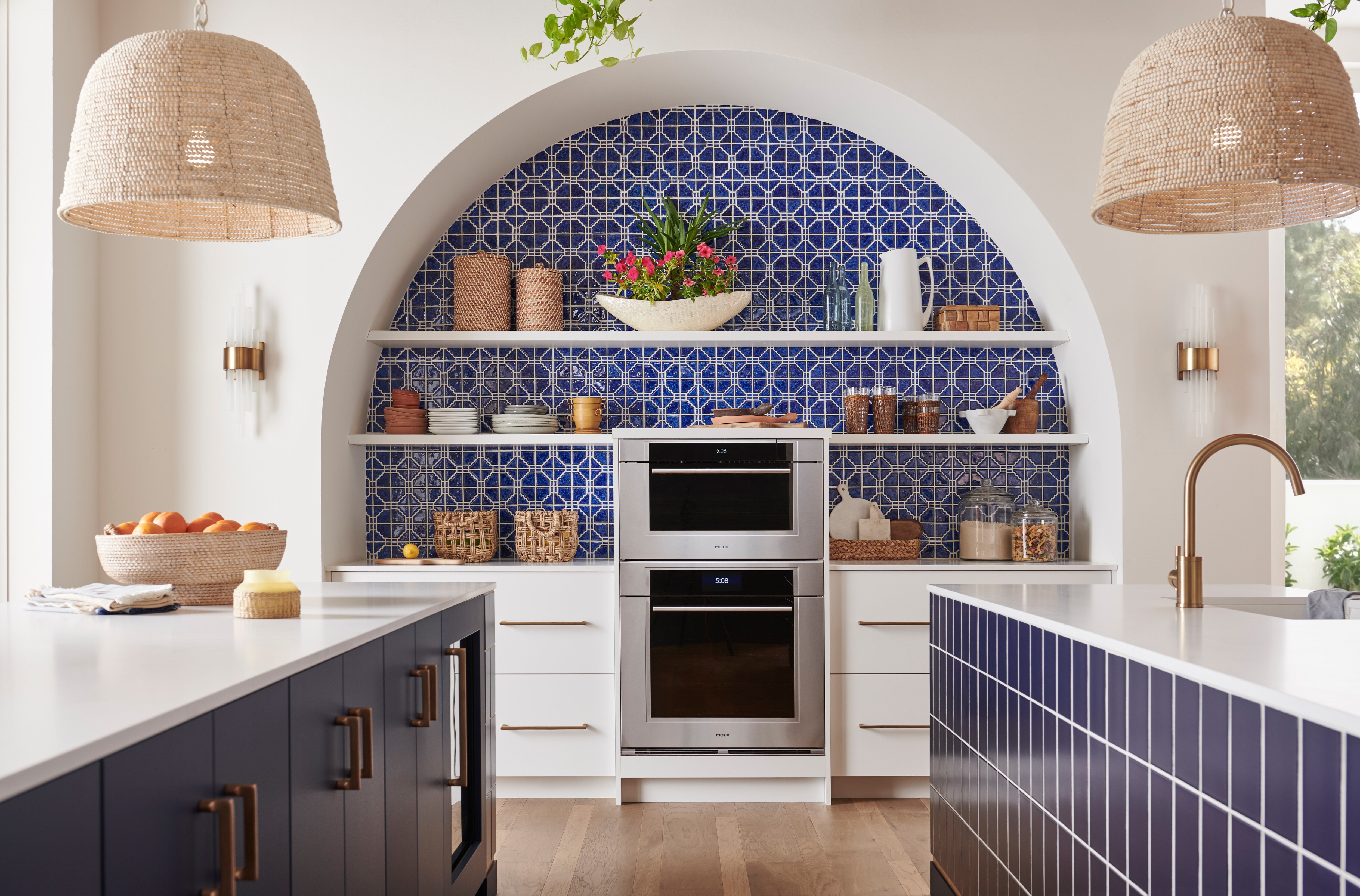 Horno de vapor con convección transicional Wolf de la serie M de 30" y horno simple empotrado transicional Wolf de la serie M de 30" mostrados juntos en un diseño de cocina mediterránea personalizada con azulejos azules, estantes abiertos y accesorios dorados