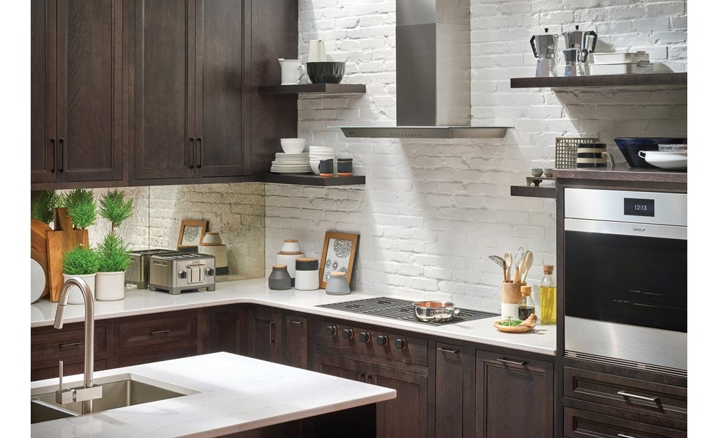 La campana extractora de placa de cocción de pared de 36" Wolf de vidrio (VW36G) exhibida contra ladrillos blancos a la vista y estantes flotantes en un diseño de cocina de lujo