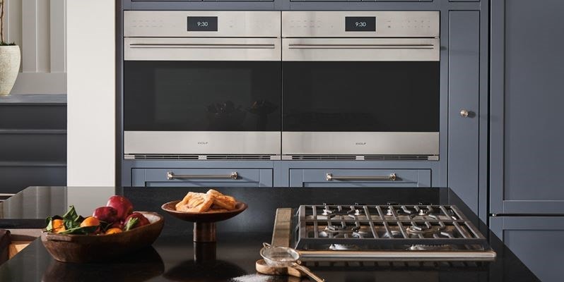 Wolf E Series (SO3050TE) Smart Wall Ovens installed side-by-side in an elegant cottage kitchen design featuring grey cabinetry with silver accent hardware.