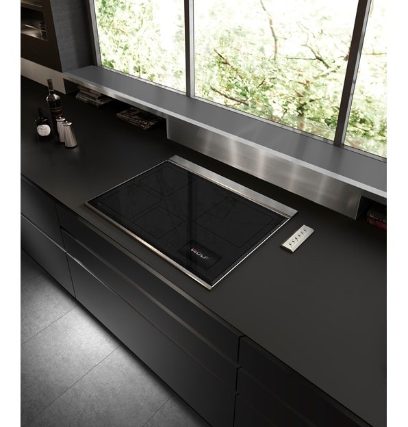 Placa de inducción transicional de 30" Wolf (CI30460T/S) exhibida en un elegante diseño de cocina ultramoderno con mostradores negros lisos, salpicadero de acero inoxidable y gabinetes sin manija 