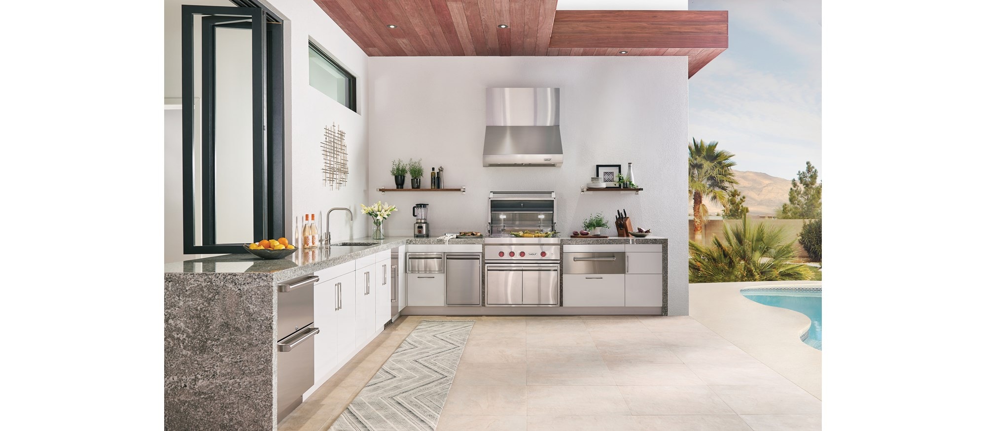 La parrilla de gas para exteriores de 36" Wolf (OG36) exhibida con el cajón calentador para exteriores de 30" Wolf (WWD30O) en esta lujosa cocina al aire libre junto a la piscina.