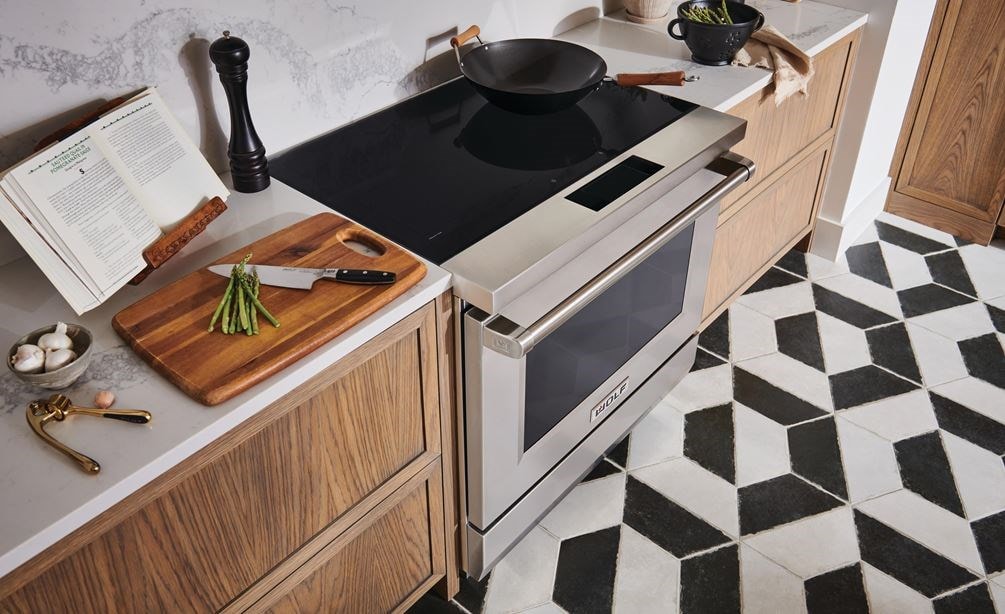 Estufa de inducción profesional de 36" y 34" Wolf (IR36550/S/P) con una sartén de wok en su superficie de cerámica negra junto a la tabla de cortar con espárragos.