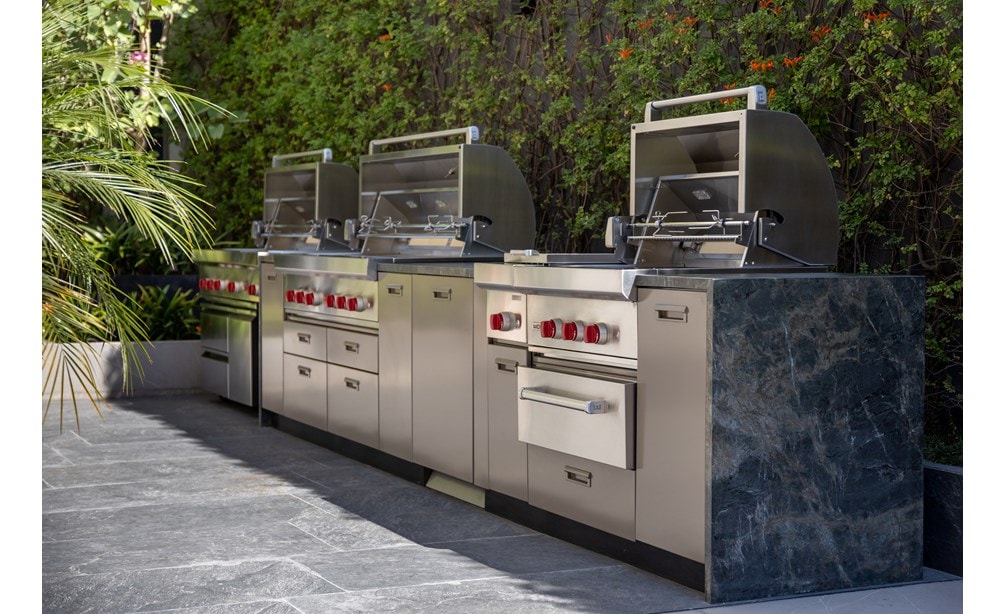Three outdoor grills side-by-side a part of Wolf Appliances in front of a brush background.